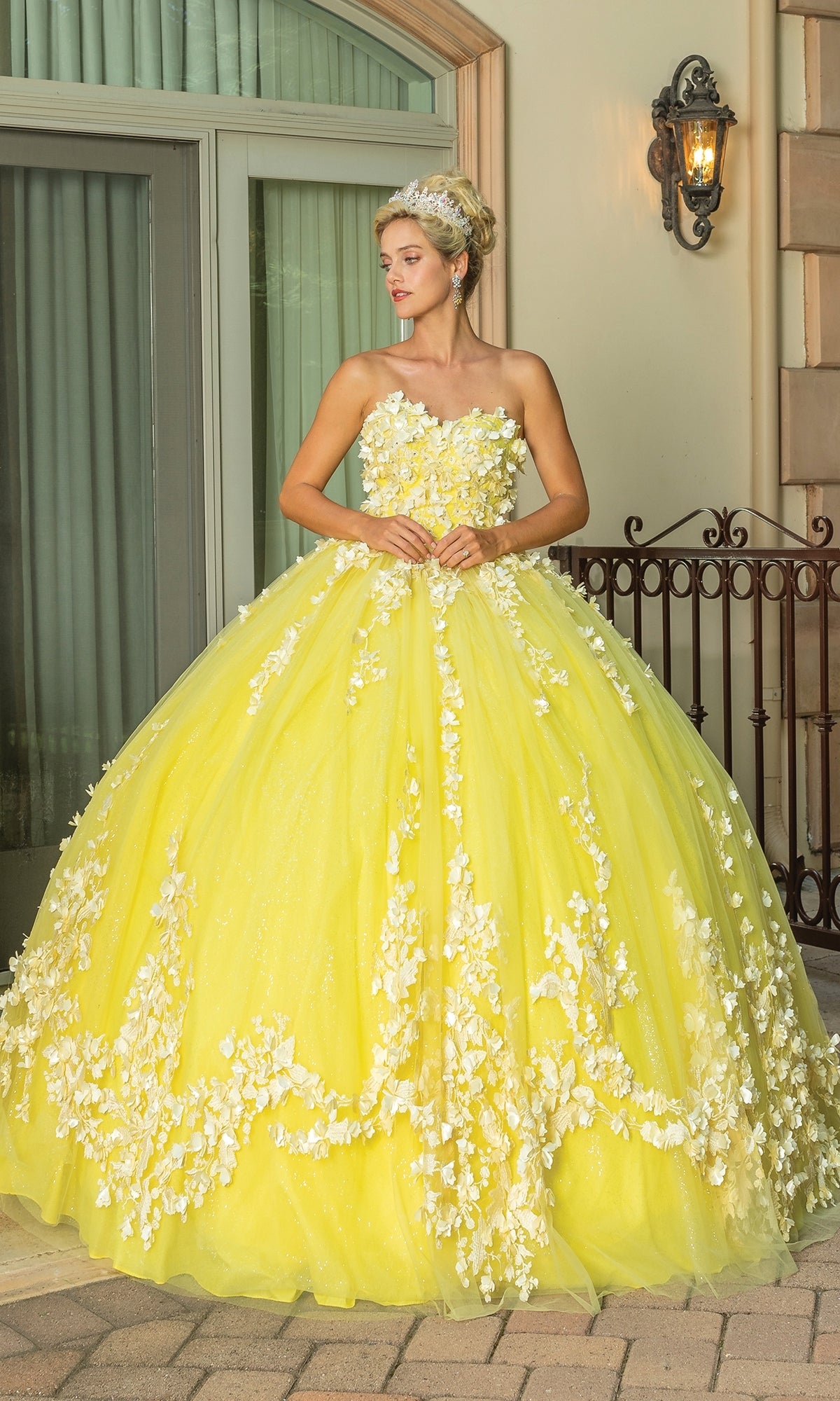 A woman stands outdoors wearing the Quinceanera Dress 1725 By Dancing Queen—a yellow a-line gown with floral appliqués, a strapless sweetheart neckline, voluminous skirt, headband accessory, and a removable cape for added flair.
